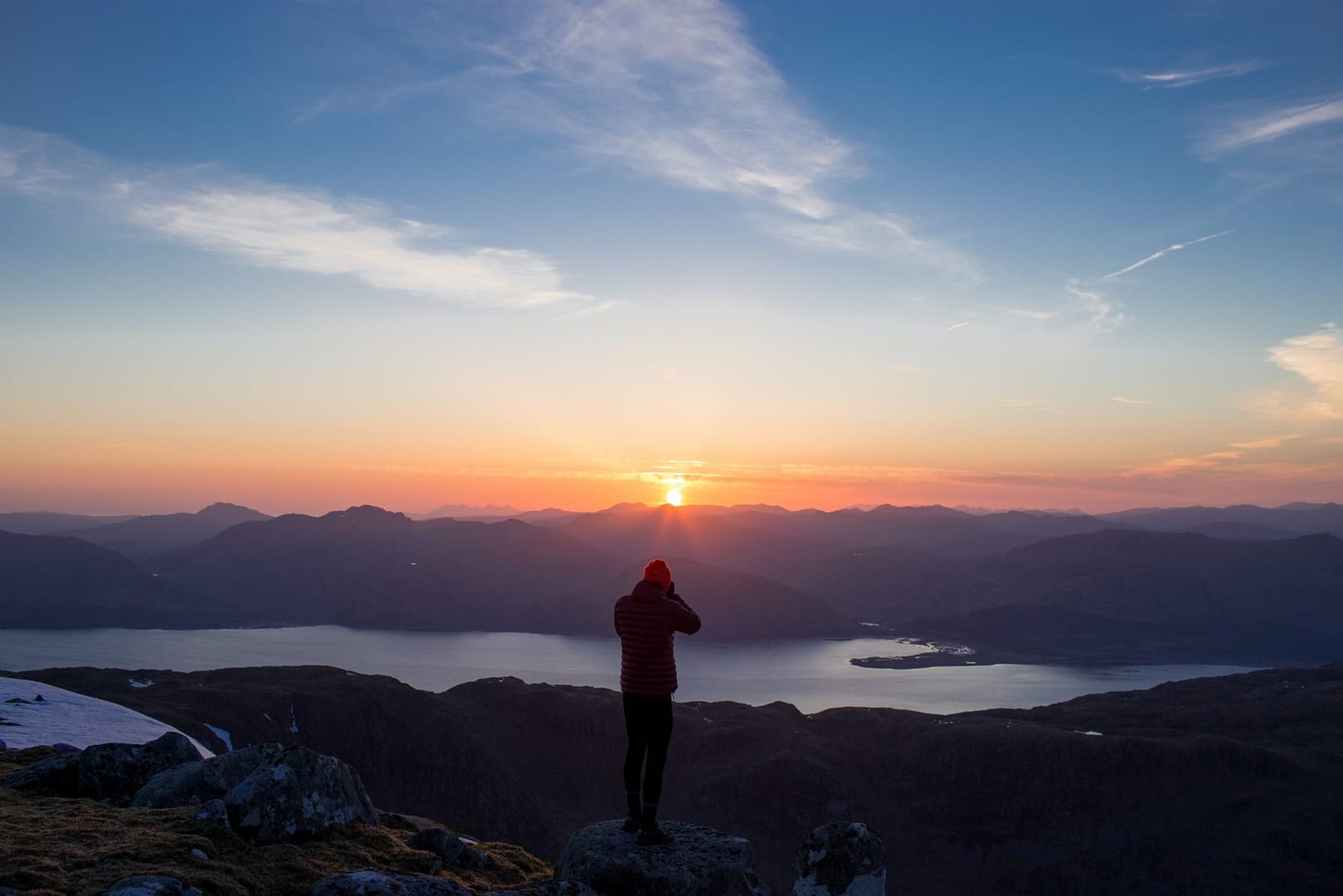 person standing on mountain ridge over lake under cloudy sky at sunset